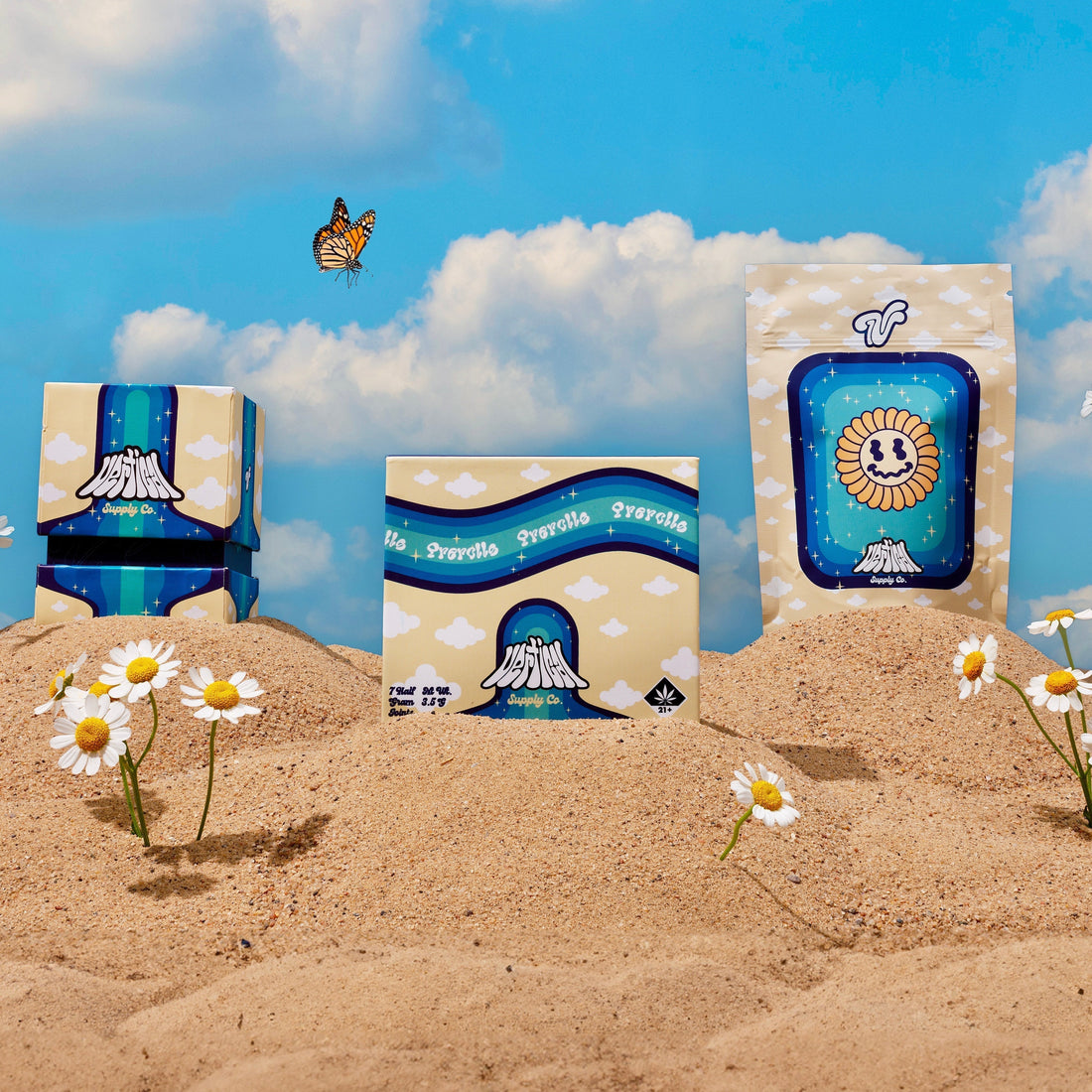 Custom packaging designs created for Vertical Supply Co. displayed in a desert sand setting with daisies and a butterfly, featuring branded boxes and a pouch with playful cloud, volcano, and smiling sun illustrations under a bright blue sky.
