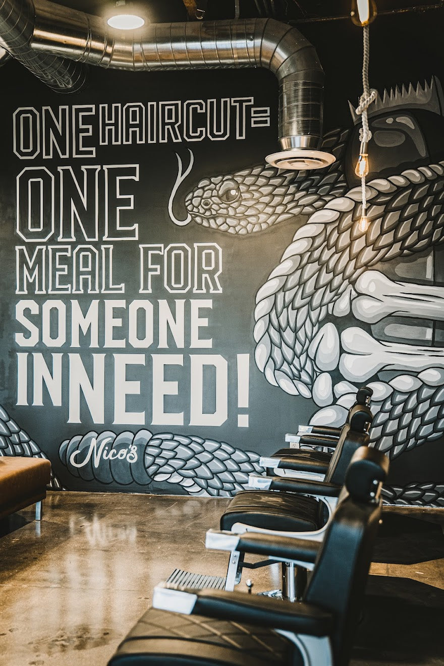 Large hand-painted mural at Nico’s Barbershop with bold lettering reading “One Haircut = One Meal for Someone in Need,” accented by a detailed grayscale snake illustration wrapping around the text.